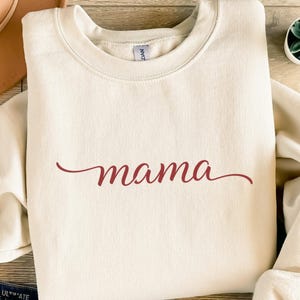 Puede incluir: Sudadera color crema con la palabra "mama" en fuente cursiva burdeos. La sudadera tiene cuello redondo y mangas largas. La tela parece suave y cómoda.