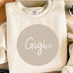 Könnte beinhalten: Cremefarbenes Sweatshirt mit dem Namen "Gigi" in weißer Schrift in einem taupefarbenen Kreis. Das Sweatshirt besteht aus einem weichen Material und hat einen Rundhalsausschnitt. Das Design ist auf der Vorderseite zentriert.
