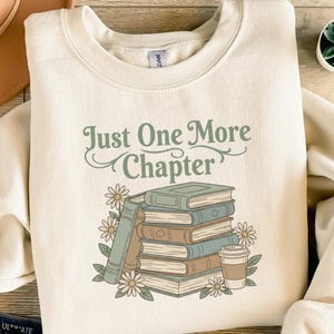 Könnte beinhalten: Cremefarbenes Sweatshirt mit dem Text "Just One More Chapter" in grüner Schrift. Die Grafik zeigt einen Stapel Bücher, Gänseblümchen und eine Kaffeetasse. Ein gemütliches, buchthematisches Design.