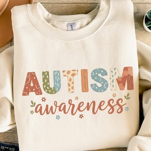 Könnte beinhalten: Cremefarbenes Sweatshirt mit den Worten "AUTISM awareness" in bunten, gemusterten Buchstaben. Das Wort "AUTISM" ist in einer Mischung aus Orange, Grün, Blau und Braun gehalten. Das Wort "awareness" ist in einer kursiven, rötlich-orangen Schrift.