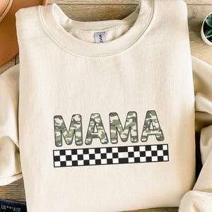 Könnte beinhalten: Cremefarbenes Sweatshirt mit dem Wort "MAMA" in einem Tarnmuster. Unter dem Wort befindet sich ein schwarz-weißes Schachbrettmuster. Das Sweatshirt ist ein lässiger Stil.