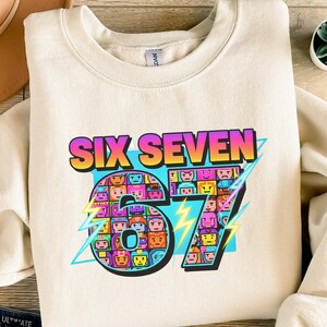 Könnte beinhalten: Cremefarbenes Sweatshirt mit dem Text "SIX SEVEN" in einer farbenfrohen Retro-Schrift. Die Zahl "67" ist mit pixeligen Gesichtern und Blitzakzenten gestaltet. Ein lässiges Unisex-Kleidungsstück.
