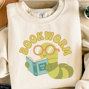 Op de afbeelding: Crèmekleurige sweatshirt met een cartoon boekenwurm die een boek leest. De worm is groen en geel, draagt een oranje bril en houdt een blauw boek vast. Het woord "BOOKWORM" staat in bijpassend oranje boven de worm.