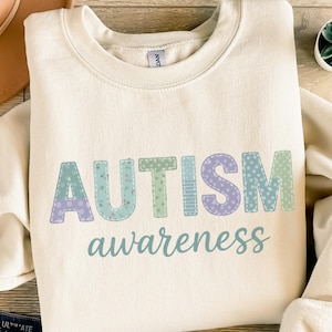 Könnte beinhalten: Cremefarbenes Sweatshirt mit dem Schriftzug "AUTISM awareness" in einem pastellfarbenen Patchwork-Design. Die Buchstaben sind in Blau-, Grün- und Lilatönen gehalten und haben einen genähten Effekt. Der Text ist in einer weichen, abgerundeten Schriftart.