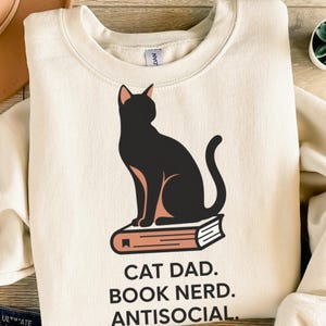 Könnte beinhalten: Ein cremefarbenes Sweatshirt mit Rundhalsausschnitt und einer Grafik einer schwarzen Katze, die auf einem pfirsichfarbenen Buch sitzt. Unter der Abbildung steht der Text: CAT DAD. BOOK NERD. ANTISOCIAL. Das Kleidungsstück hat einen gerippten Kragen.