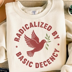 Op de afbeelding: Crèmekleurige sweatshirt met een ronde halslijn. De voorkant heeft een cirkelvormig ontwerp met de woorden "RADICALIZED BY BASIC DECENCY" in een roestkleurig lettertype. Een roestkleurige duif met een olijftak staat in het midden van het ontwerp.