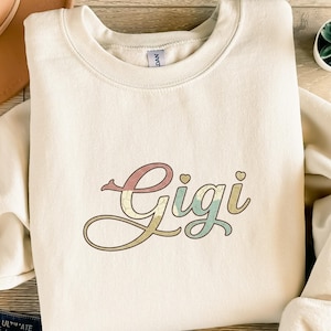 Könnte beinhalten: Cremefarbenes Sweatshirt mit dem Namen "Gigi" in einem Retro-Schriftzug. Der Text ist in Rosa-, Blau- und Gelbtönen gehalten. Das Sweatshirt hat einen Rundhalsausschnitt und lange Ärmel. Das Wort "Gigi" ist der Fokus des Designs.