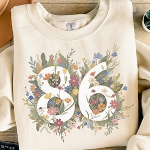 Puede incluir: Sudadera color crema con el número 86 en blanco, rodeada de un vibrante arreglo floral. Las flores incluyen flores rosas, amarillas, azules y naranjas, con follaje verde. La sudadera tiene cuello redondo y mangas largas.
