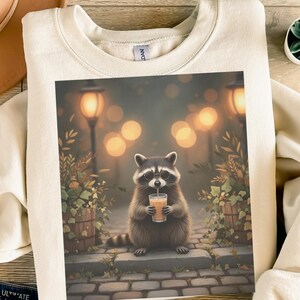 Könnte beinhalten: Cremefarbenes Sweatshirt mit einem Waschbären, der eine Brille trägt und ein Getränk mit einem Strohhalm hält. Der Waschbär ist vor dem Hintergrund einer Straßenszene mit weichen, verschwommenen Lichtern und Grün angeordnet. Das Sweatshirt hat eine neutrale Farbe.