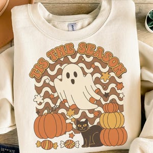 Könnte beinhalten: Cremefarbenes Sweatshirt mit einem Retro-Halloween-Design. Die Grafik zeigt einen weißen Geist, Kürbisse, eine schwarze Katze und den Text "TIS THE SEASON" in Orange und Braun. Herbstblätter und Süßigkeiten vervollständigen das festliche Design.