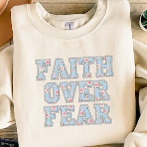 Könnte beinhalten: Cremefarbenes Sweatshirt mit dem Text "FAITH OVER FEAR" in hellblauen Buchstaben, gefüllt mit einem rosa Blumenmuster. Die Buchstaben haben eine silberne Umrandung, die ein dekoratives Design erzeugt.