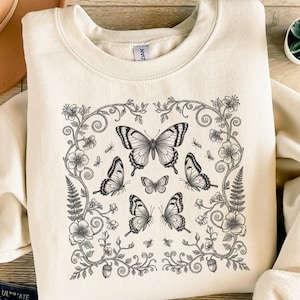 Könnte beinhalten: Cremefarbenes Sweatshirt mit einem detaillierten schwarz-weißen Schmetterlingsdesign. Das Design umfasst mehrere Schmetterlinge, Blumen und Blätter in einem quadratischen Rahmen. Das Sweatshirt hat einen Rundhalsausschnitt und lange Ärmel.