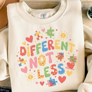 Könnte beinhalten: Cremefarbenes Sweatshirt mit einem farbenfrohen Aufdruck mit der Aufschrift "DIFFERENT NOT LESS". Das Design umfasst Puzzleteile, Herzen und Sonnen in verschiedenen Farben. Das Sweatshirt hat einen Rundhalsausschnitt und lange Ärmel.