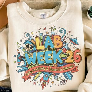 Könnte beinhalten: Cremefarbenes Sweatshirt mit farbenfrohem Grafikdesign. Das Design zeigt die Worte "LAB WEEK 26" in großen, umrandeten Buchstaben sowie wissenschaftliche Illustrationen wie Bechergläser, ein Mikroskop und eine Rakete. Der Text "Going to Infinity & Byond for Patients!" ist ebenfalls enthalten.