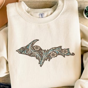 Könnte beinhalten: Cremefarbenes Sweatshirt mit einer detaillierten Grafik einer Staatsumrisslinie, verziert mit floralen und Rankenmustern in Braun, Türkis und Creme. Das Design befindet sich mittig auf der Vorderseite des langärmeligen Kleidungsstücks.