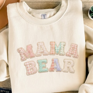 Könnte beinhalten: Cremefarbenes Sweatshirt mit den Worten "MAMA BEAR" in pastellfarbenen Patchwork-Buchstaben. Die Buchstaben haben Blumen-, Vichy- und Tupfenmuster. Das Sweatshirt hat einen Rundhalsausschnitt und lange Ärmel.