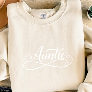 Könnte beinhalten: Cremefarbenes Sweatshirt mit dem Wort "Auntie" in eleganter weißer Schrift. Das Wort ist mittig auf der Vorderseite des Sweatshirts platziert, mit einem kleinen Herzdetail. Das Sweatshirt hat einen Rundhalsausschnitt und lange Ärmel.