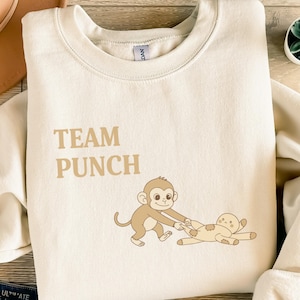 Puede incluir: Sudadera de color crema con el texto "TEAM PUNCH" en una fuente marrón claro. El gráfico presenta un mono de dibujos animados tirando de una muñeca. La sudadera tiene cuello redondo y mangas largas.