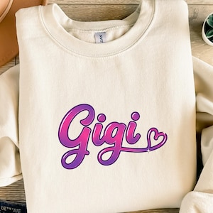 Könnte beinhalten: Cremefarbenes Sweatshirt mit dem Namen "Gigi" in einem rosa-violetten Farbverlauf. Das Wort endet mit einem Herzen. Das Sweatshirt hat einen lässigen Stil.