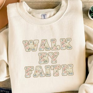 Könnte beinhalten: Cremefarbenes Sweatshirt mit dem Schriftzug "WALK BY FAITH" in floralen Buchstaben. Die Buchstaben haben eine hellbraune Umrandung und zeigen rosa, blaue und cremefarbene Blumen. Das Sweatshirt hat einen Rundhalsausschnitt und lange Ärmel.