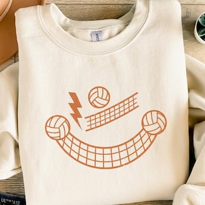 Könnte beinhalten: Cremefarbenes Sweatshirt mit einem Volleyball-Motiv. Das Design zeigt ein lächelndes Gesicht aus Volleybällen, einem Netz und einem Blitz in Orange- und Brauntönen. Das Sweatshirt ist aus einem weichen Material gefertigt.