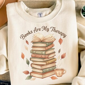 Könnte beinhalten: Cremefarbenes Sweatshirt mit dem Text "Books Are My Therapy" in brauner Schrift. Die Grafik zeigt einen Stapel Bücher, ein offenes Buch, Herbstblätter und eine Kaffeetasse. Das Design ist auf der Vorderseite aufgedruckt.