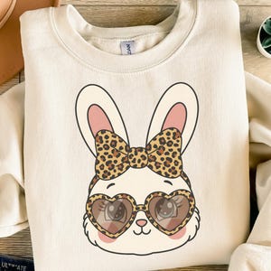 Könnte beinhalten: Cremefarbenes Sweatshirt mit einem Cartoon-Hasengesicht. Der Hase hat herzförmige Sonnenbrillen und eine Schleife mit Leopardenmuster. Das Design umfasst rosa Innenohren und rosige Wangen. Das Bild ist auf der Vorderseite zentriert.