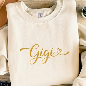 Könnte beinhalten: Cremefarbenes Sweatshirt mit dem Namen "Gigi" in goldfarbener Glitzerschrift. Das Wort "Gigi" ist in einer Schreibschrift mit einem Herzsymbol am Ende geschrieben. Das Sweatshirt hat einen Rundhalsausschnitt und lange Ärmel.