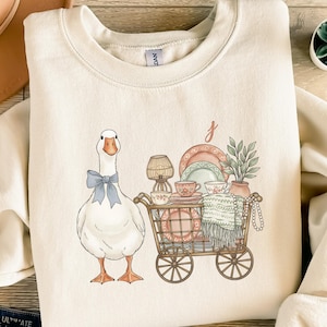Könnte beinhalten: Cremefarbenes Sweatshirt mit einer Gans, die eine blaue Fliege trägt. Ein Einkaufswagen mit Geschirr, Tassen und einer Decke steht neben der Gans. Der Wagen hat eine Lampe, Pflanzen und eine Perlenkette. Der Buchstabe "J" befindet sich über dem Wagen.