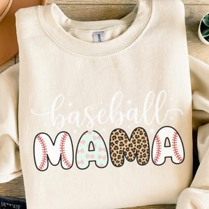 Könnte beinhalten: Cremefarbenes Sweatshirt mit den Worten "baseball MAMA" in Weiß und verschiedenen Farben. Das Wort "baseball" ist kursiv geschrieben, und das Wort "MAMA" hat Baseball- und Leopardenmuster.