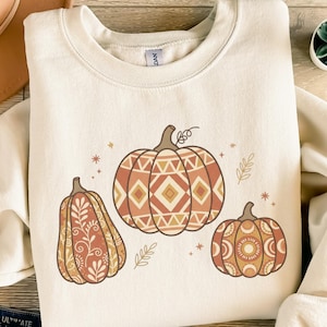 Könnte beinhalten: Cremefarbenes Sweatshirt mit drei dekorativen Kürbissen in Orange-, Braun- und Beigetönen. Die Kürbisse haben aufwendige geometrische und florale Designs. Ein Bekleidungsstück zum Thema Herbst.
