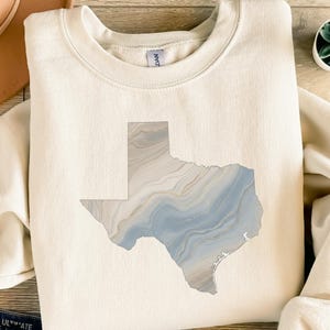 Könnte beinhalten: Cremefarbenes Sweatshirt mit einem marmorierten Umriss des Bundesstaates Texas in Blau-, Grau- und Beigetönen. Das Design befindet sich mittig auf der Vorderseite des langärmeligen Rundhals-Oberteils. Das Sweatshirt ist aus weichem Material.