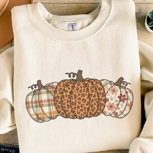 Könnte beinhalten: Cremefarbenes Sweatshirt mit Rundhalsausschnitt und drei Kürbissen mit verschiedenen Mustern: Karomuster, Leopardenmuster und Blumenmuster. Das langärmlige Oberteil ist ideal für den Herbst.
