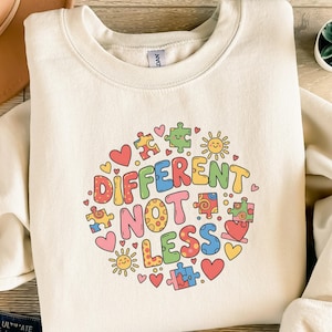 Könnte beinhalten: Cremefarbenes Sweatshirt mit einem farbenfrohen Aufdruck mit der Aufschrift "DIFFERENT NOT LESS". Das Design umfasst Puzzleteile, Herzen und Sonnen in verschiedenen Farben. Das Sweatshirt hat einen Rundhalsausschnitt und lange Ärmel.