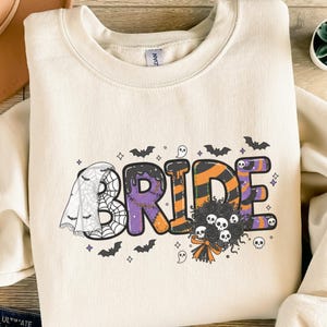 Op de afbeelding: Crèmekleurige sweatshirt met het woord "BRIDE" in een Halloween-thema. De letters zijn paars, oranje en zwart, met een spinnenweb en vleermuizen. Een boeket van schedels en een sluier maken de spookachtige look compleet.