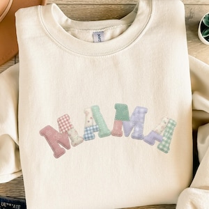 Könnte beinhalten: Cremefarbenes Sweatshirt mit dem Wort "MAMA" auf der Vorderseite. Die Buchstaben sind im Patchwork-Stil gehalten und verwenden verschiedene pastellfarbene Stoffe mit Blumen- und Vichy-Mustern. Das Sweatshirt hat einen Rundhalsausschnitt.