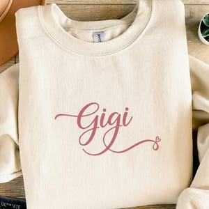 Könnte beinhalten: Cremefarbenes Sweatshirt mit dem Namen "Gigi" in einer rosafarbenen Schreibschrift. Das Design beinhaltet ein kleines Herz am Ende des Namens. Das Sweatshirt hat einen Rundhalsausschnitt und lange Ärmel.