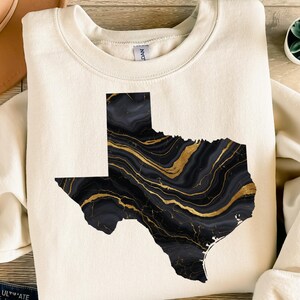 Op de afbeelding: Crèmekleurige sweatshirt met een zwart en goud gemarmerde omtrek van de staat Texas. Het ontwerp heeft een vloeiend, abstract patroon met gouden aderen tegen een donkere achtergrond. De sweatshirt is een casual, comfortabele stijl.