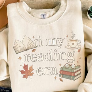 Könnte beinhalten: Cremefarbenes Sweatshirt mit dem Schriftzug "in my reading era" in einer verspielten Schriftart. Das Design umfasst ein offenes Buch, eine Tasse Tee, ein Ahornblatt, eine Brille und gestapelte Bücher. Ein gemütliches Kleidungsstück zum Thema Lesen.