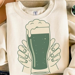 Könnte beinhalten: Cremefarbenes Sweatshirt mit einem grünen Bier-Motiv. Das Design zeigt ein Bierglas mit Schaum, gehalten von zwei Händen. Die Grafik ist im einfachen Linienstil gehalten, ideal für Freizeitkleidung.