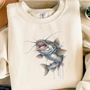 Könnte beinhalten: Cremefarbenes Sweatshirt mit einer Aquarellillustration eines Welses. Der Fisch ist in Grau- und Blautönen gehalten und hat lange Barteln und detaillierte Flossen. Blaue Aquarellfarbspritzer akzentuieren das Design. Das Sweatshirt hat einen Rundhalsausschnitt.