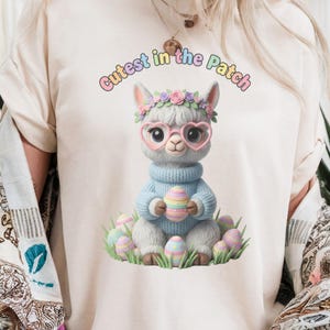Könnte beinhalten: Cremefarbenes T-Shirt mit einem Cartoon-Lama, das eine herzförmige Brille und einen Blumenkranz trägt. Das Lama hält ein pastellfarbenes Osterei, umgeben von weiteren Eiern. Der Text oben lautet "Cutest in the Patch."