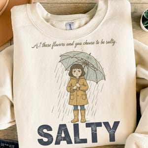 Könnte beinhalten: Cremefarbenes Sweatshirt mit einer Grafik eines Mädchens unter einem Regenschirm im Regen. Der Text "At these flavors and you choose to be salty" steht über der Grafik, das Wort "SALTY" darunter.
