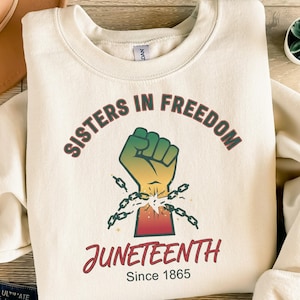 Könnte beinhalten: Cremefarbenes Sweatshirt mit dem Text "Sisters in Freedom" über einer Grafik einer Faust, die Ketten sprengt. Darunter steht das Wort "Juneteenth" in einer Schriftart, mit "Since 1865" darunter.