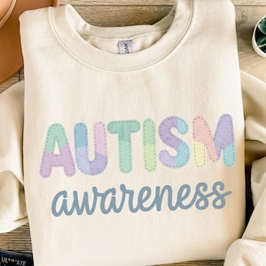Könnte beinhalten: Cremefarbenes Sweatshirt mit den Worten "AUTISM awareness" in Pastellfarben. Das Wort "AUTISM" ist in einem genähten Buchstabenstil gehalten, wobei jeder Buchstabe eine andere Farbe hat. Das Wort "awareness" ist in hellblauer Schrift geschrieben.