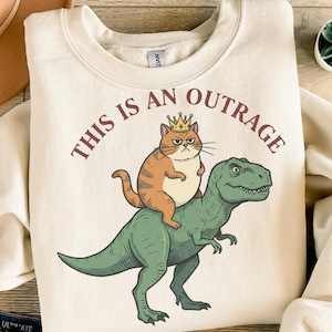 Puede incluir: Sudadera color crema con la frase "THIS IS AN OUTRAGE" en letras granates. El diseño presenta un gato naranja con corona montado sobre un dinosaurio verde. El gato tiene una expresión de enfado.
