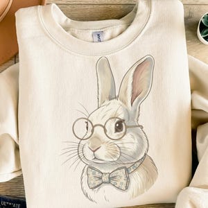 May include: A cream-coloured sweatshirt with a detailed illustration of a white rabbit wearing round glasses and a floral bow tie. The rabbit has long ears and is centered on the front. The sweatshirt has a crew neck.
