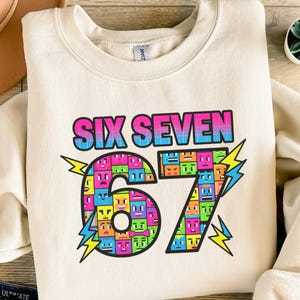 Könnte beinhalten: Cremefarbenes Sweatshirt mit dem Text "SIX SEVEN" in einem Farbverlauf aus Pink, Blau und Grün. Die Zahlen "67" sind mit bunten pixeligen Gesichtern gefüllt, mit Blitzgrafiken auf beiden Seiten.