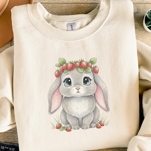 Könnte beinhalten: Cremefarbenes Sweatshirt mit einer Illustration eines niedlichen grauen Hasen mit Erdbeerkrone. Der Hase hat große blaue Augen und rosa gefütterte Ohren. Erdbeeren und grüne Blätter sind um den Hasen verstreut. Ein bezauberndes Design für einen lässigen Look.