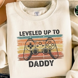 Könnte beinhalten: Cremefarbenes Sweatshirt mit dem Text "LEVELED UP TO DADDY" über einer Grafik eines Videospiel-Controllers im Retro-Stil. Das Controller-Design hat ein kreuzförmiges Steuerkreuz, Knöpfe und ein Vintage-Farbschema aus Orange, Gelb und Braun.